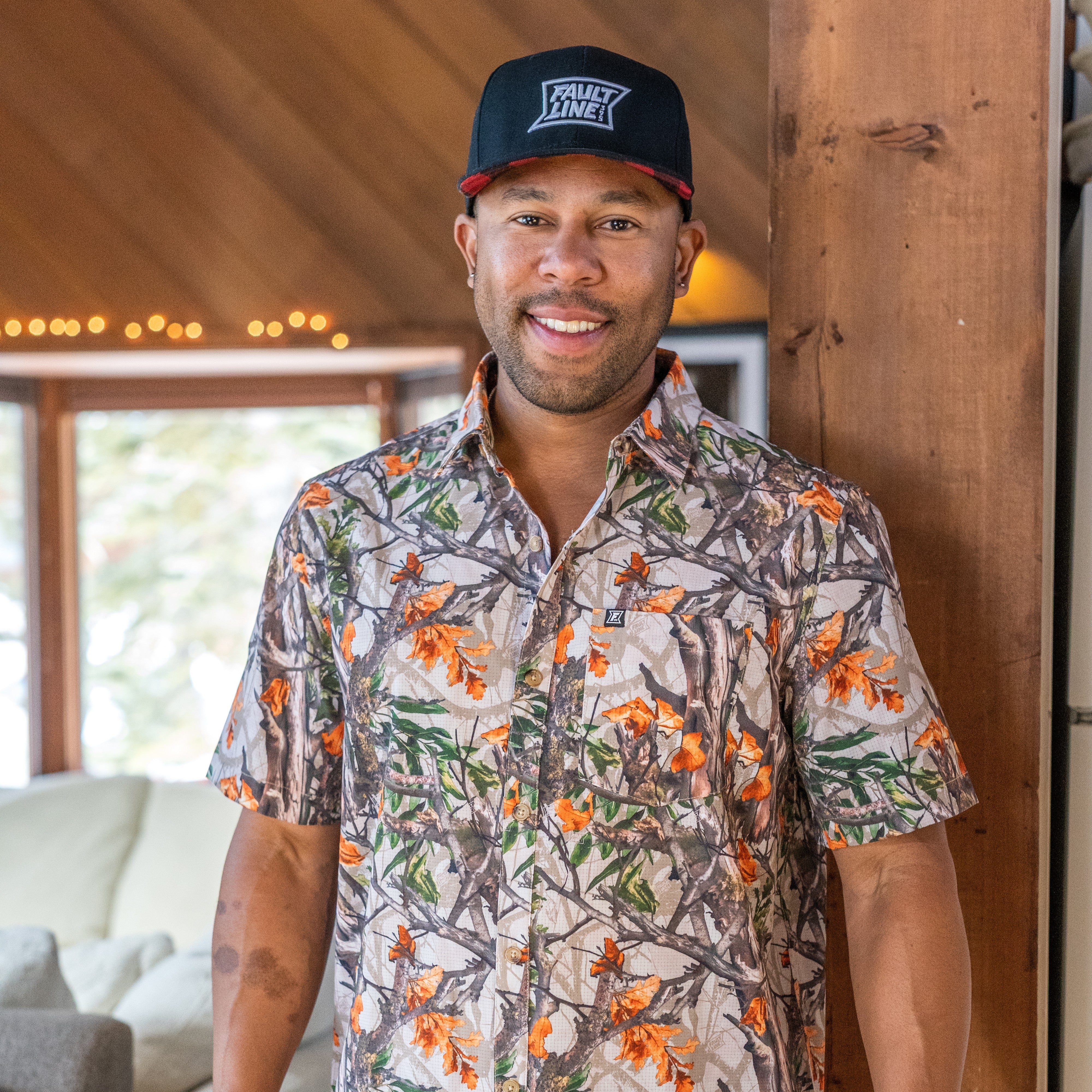 Man wearing a real tree patterned shirt and faultline395 cap inside a mountain cabin
