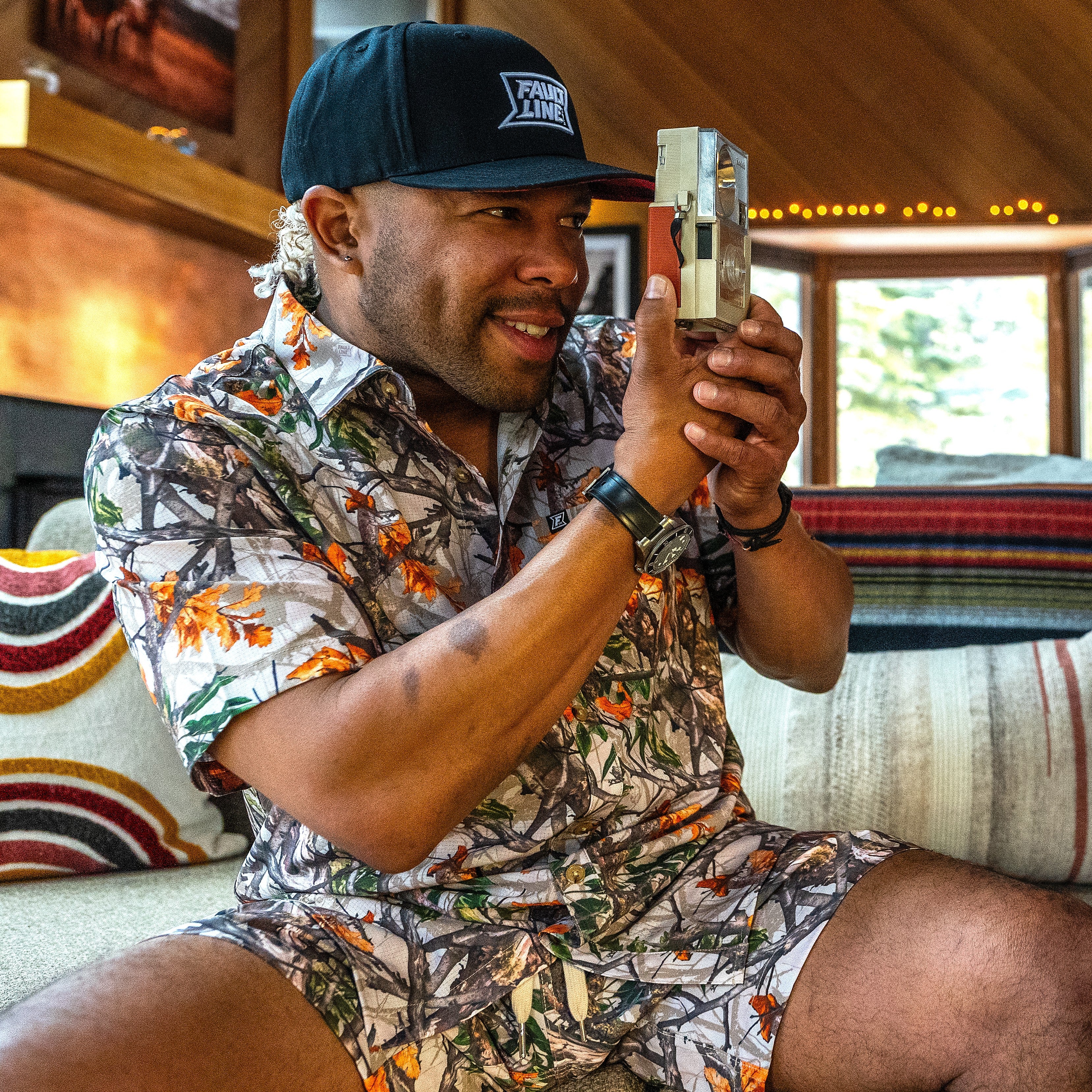 Man holding a retro camera wearing a real tree camo outfit set inside a mountain cabin